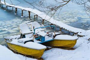 blauw en geel catamaran in een winter dag, gedekt met sneeuw staand in de buurt de bevroren rivier- en houten brug. catamaran Aan sneeuw onder de boom. detailopname foto