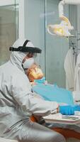 tandheelkunde dokter in beschermend pak gebruik makend van gesteriliseerd tandheelkundig gereedschap onderzoeken senior geduldig gedurende coronavirus pandemie. medisch team pratend met vrouw vervelend gezicht schild, overall, masker en handschoenen foto