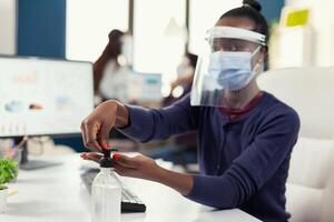 Afrikaanse ondernemer gebruik makend van hand- ontsmettingsmiddel Bij werkplaats vervelend gezicht masker. zakenvrouw in nieuw normaal werkplaats desinfecteren terwijl collega's werken in achtergrond. foto