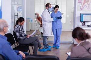 tandarts afwerking tanden behandeling voor oud Mens analyseren röntgenstraal radiografie met verpleegster in stomatologie aan het wachten kamer vol met patiënten. mensen vulling het formulier in tandheelkunde gang. foto