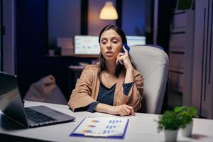 werkverslaafde manager pratend met klant over- de telefoon in de avond. vrouw ondernemer werken laat Bij nacht in zakelijke bedrijf aan het doen na een tijdje in de Cursus van telefoon telefoongesprek. foto