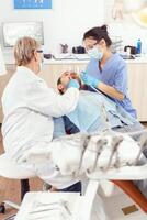 stomatoloog senior vrouw het uitvoeren van examen en nemen zorg van tanden gebruik makend van tandheelkundig hulpmiddelen. orthodontist sprekend naar ziek Mens zittend Aan stomatologisch stoel in modern kliniek foto