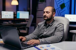 Kaukasisch ondernemer werken Aan deadline in donker kantoor typen Aan laptop. slim zakenman zittend Bij zijn werkplaats in de Cursus van laat nacht uren aan het doen zijn functie. foto