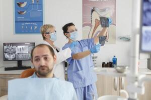 portret van geduldig met kiespijn aan het wachten voor medisch procedure gedurende stomatologisch overleg in tandheelkunde kantoor kamer. in achtergrond medisch team analyseren tanden radiografie. geneeskunde onderhoud foto