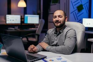 portret van freelancer op zoek Bij camera zittend Bij bureau met grafieken. slim zakenman zittend Bij zijn werkplaats in de Cursus van laat nacht uren aan het doen zijn functie. foto