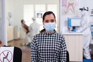 portret van vrouw in tandheelkundig kantoor op zoek Aan camera vervelend gezicht masker zittend Aan stoel in aan het wachten kamer kliniek terwijl dokter werken. concept van nieuw normaal tandarts bezoek in coronavirus uitbraak. foto