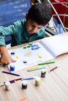 slim Indisch weinig jongen uitvoeren duim schilderij met verschillend kleurrijk water kleur uitrusting gedurende de zomer vakanties, schattig Indisch kind aan het doen kleurrijk duim schilderij tekening Aan houten tafel foto