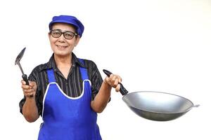 Aziatisch senior vrouw, draagt blauw schort en pet, houdt frituren pan en spatel of pollepel. kijken Bij camera, glimlacht, voelt zelfverzekerd geïsoleerd Aan wit achtergrond. concept, liefde Koken. keuken levensstijl. foto