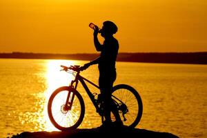 silhouet van fietser. sportief vent wielersport in helm drankjes water Aan de natuurlijk achtergrond van instelling de zon in de avond foto