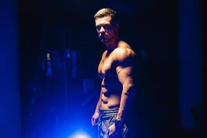 aantrekkelijk bodybuilder poseren en tonen uit spieren Aan donker achtergrond. voor de helft beurt naar de camera. detailopname. blauw licht filter. sterk atleet met naakt torso. geschiktheid concept. foto