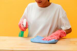 schoonmaak concept. Aziatisch vrouw Holding een verstuiven fles met schoonmaakster en een controleren kleding naar schoon een geel achtergrond. huisvrouw wie reinigt de huis. gelukkig schoonmaak foto