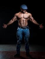 elegant bodybuilder staand Leuk vinden ster Bij studio. sexy gespierd Mens in blauw hoed poseren voor camera. foto