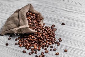 zak van natuurlijk bruin koffie bonen en sommige verspreide zaden Aan de licht houten achtergrond. geroosterd koffie granen morsen uit van zak Aan de tafel. foto