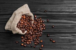 jute- zak vol van geroosterd koffie bonen en verspreide Aan een houten tafel foto