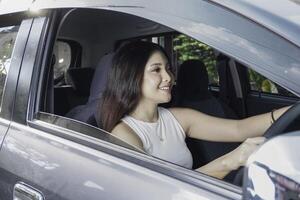 aantrekkelijk jong Aziatisch vrouw in gewoontjes slijtage is glimlachen terwijl het rijden een auto foto