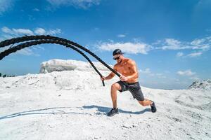 sterk bodybuilder in een pet en zonnebril opleiding met touwen buitenshuis. zwaar opleiding in een groeve. wit landschap. foto