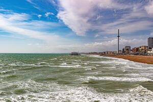 zonnig strandlandschap met golven, pier in afstand, en bewolkt blauw lucht foto