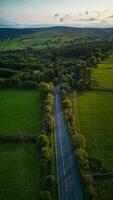 leeg land weg meanderend door weelderig groen velden met bebost heuvels in de achtergrond gedurende zonsondergang in yorkshire. foto