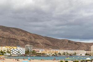 toneel- visie van een aan het strand met toeristen en een berg backdrop onder bewolkt luchten in los Cristianos, tenerife. foto
