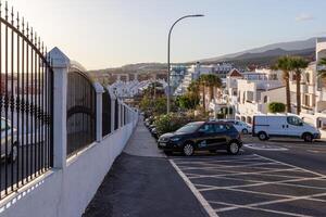 stil woon- straat met geparkeerd auto's en wit huizen, metaal hek Aan de links, en heuvels in de achtergrond Bij schemer in los Cristianos, tenerife. foto