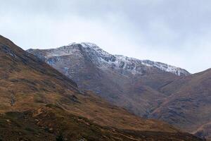 majestueus berg landschap met met sneeuw bedekt top en herfst- bruin heuvels onder een bewolkt lucht in Schotland. foto