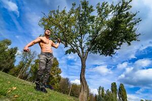 sterk gespierd bodybuilder atleet Mens poseren en pompen omhoog spieren Aan een groen boom achtergrond. gezond sport leven concept. visie van hieronder foto