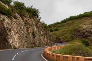 golvend berg weg met beschermend belemmeringen, omringd door rotsachtig terrein en groen onder een bewolkt lucht berg landschap met blauw lucht en wolken in tenerife. foto