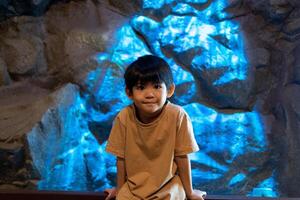 weinig Aziatisch jongen zit tegen een rots achtergrond, blauw lichten spatten de achtergrond. foto