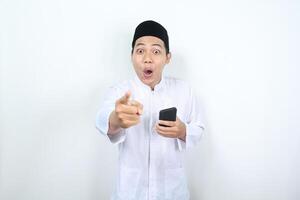 versteld staan Aziatisch moslim Mens richten vinger naar camera met Holding smartphone geïsoleerd foto