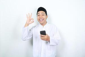 glimlachen Aziatisch moslim Mens tonen Oke tekens met Holding mobiel telefoon geïsoleerd foto