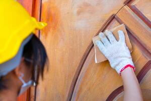 dichtbij omhoog hand- vrouw arbeider vervelend een veiligheid helm gebruik makend van schuurpapier naar Pools een houten deur Bij huis. foto