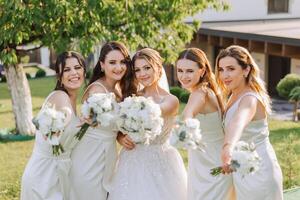 een brunette bruid en haar bruidsmeisjes in identiek wit jurken zijn staand Aan de achtergrond van natuur met boeketten van bloemen in hun handen. meisjes in identiek jurken zijn maken uit Bij een bruiloft foto