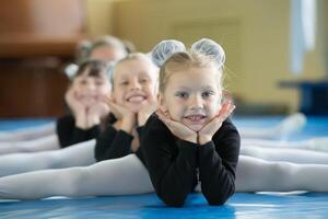 weinig meisjes gymnasten zitten Aan de splitst. kinderen Doen fysiek oefening. jong ballerina's.. opleiding van de eerste groep Aan gymnastiek.acrobatiek les foto