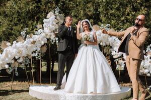 bruiloft ceremonie in natuur. de bruid en bruidegom in de buurt de bloem boog. meester van ceremonies in donker bril Bij een bruiloft gedurende een prestatie tegen de achtergrond van de bruid en bruidegom. foto
