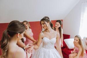 bruiloft ochtend. bruidsmeisjes helpen zetten Aan de wit bruiloft jurk. een jong vrouw is voorbereidingen treffen naar ontmoeten haar bruidegom en hebben pret met haar vrienden foto