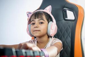 portret van een weinig meisje met pc gamer accessoires. ze looks Bij de camera. ze draagt hoofdtelefoons met kat oren en is zittend in een gaming stoel in voorkant van de toetsenbord. foto