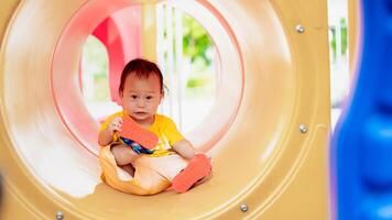 Aziatisch glimlachen weinig baby jongen geniet speeltijd in de park gedurende een zonnig zomer dag. kleuter kind zit en kruipt in de Oranje Geel speelgoed- tunnel Bij de speelplaats. weinig kind oud een jaar oud. foto