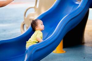 Aziatisch baby jongen klimt de kinderen glijbaan Aan speelplaats. kind spelen gelukkig in zomer of voorjaar keer. kleuter spelen buitenshuis. kind Speel Aan school- of kleuterschool werf.actief zoon Aan kleurrijk glijbaan foto