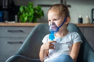 schattig weinig meisje zijn zittend en Holding een vernevelaar masker leunend tegen de gezicht Bij huis Aan ziek vertrekken, luchtweg behandeling concept foto