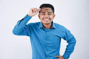 glimlachen of gelukkig knap Aziatisch zakenman hijs- bril naar voorhoofd met hand- op zoek camera en vervelend blauw overhemd geïsoleerd Aan wit achtergrond foto