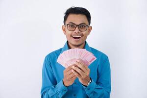 verrast of geschokt knap jong Aziatisch zakenman in vervelend blauw overhemd en bril Holding ventilator van Indonesië bankbiljetten in handen geïsoleerd Aan wit achtergrond foto