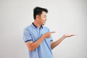 glimlachen Aziatisch Mens met een Open hand- met vingers richten naar de kant vervelend blauw t overhemd geïsoleerd Aan wit achtergrond foto