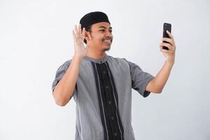 portret van knap jong Aziatisch moslim Mens vervelend koko kleren en gebruik makend van smartphone nemen een video telefoontje en golvend glimlach Bij telefoon geïsoleerd Aan wit achtergrond foto