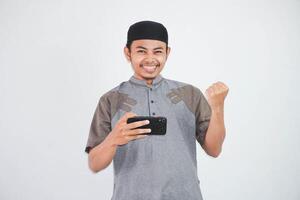 opgewonden of gelukkig jong Aziatisch moslim Mens Holding Aan mobiel telefoon en handen in vuisten aan het doen winnaar gebaar vervelend koko kleren geïsoleerd over- wit achtergrond foto