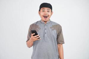 geschokt jong Aziatisch moslim Mens Holding mobiel telefoon vervelend moslim kleren geïsoleerd Aan wit achtergrond foto