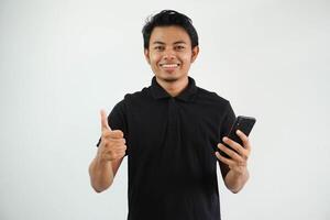 glimlachen jong Aziatisch Mens in zwart polo t overhemd Holding mobiel telefoon en maken duim omhoog gebaar, goedkeuren mooi zo ding geïsoleerd Aan wit achtergrond foto