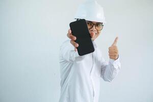 jong Aziatisch architect Mens vervelend bouwer veiligheid helm over- geïsoleerd achtergrond aan het doen gelukkig duimen omhoog gebaar en Holding mobiel telefoon met hand. foto