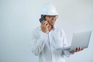 jong Aziatisch mannetje ingenieur vervelend wit moeilijk hoed Holding laptop en Aan telefoontje voor bouw werk geïsoleerd Aan wit achtergrond, kopiëren ruimte. foto