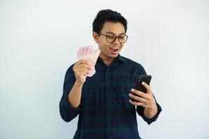 jong Aziatisch Mens tonen verrast uitdrukking wanneer op zoek naar zijn mobiel telefoon terwijl Holding papier geld geïsoleerd Aan wit achtergrond. foto