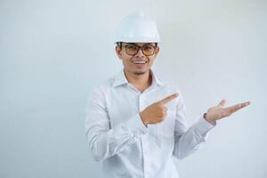 jong Aziatisch architect Mens vervelend wit moeilijk hoed veiligheid helm op zoek camera met vinger richten naar zijn Open hand- palm geïsoleerd Aan wit achtergrond foto